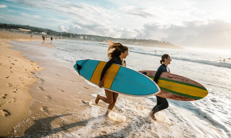 Zwei junge Frauen laufen mit Surfbrettern in die Brandung der North Coast.