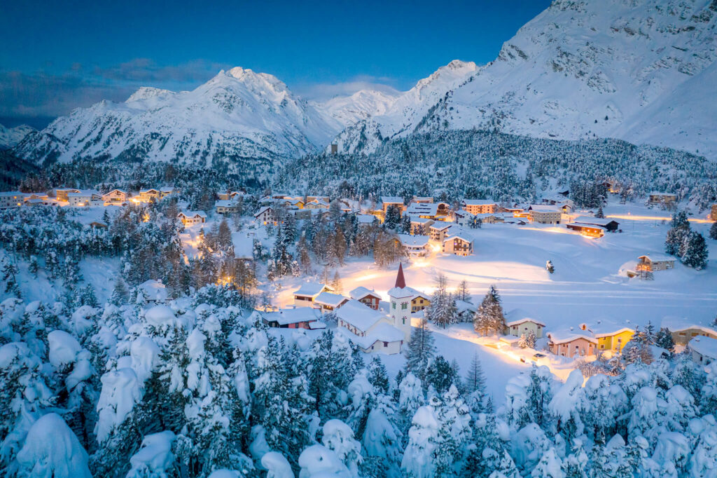 Idyllisches Schweizer Dorf mit schwach gelb erleuchteten Fenstern, umgeben von schneebedeckten Alpenbergen.