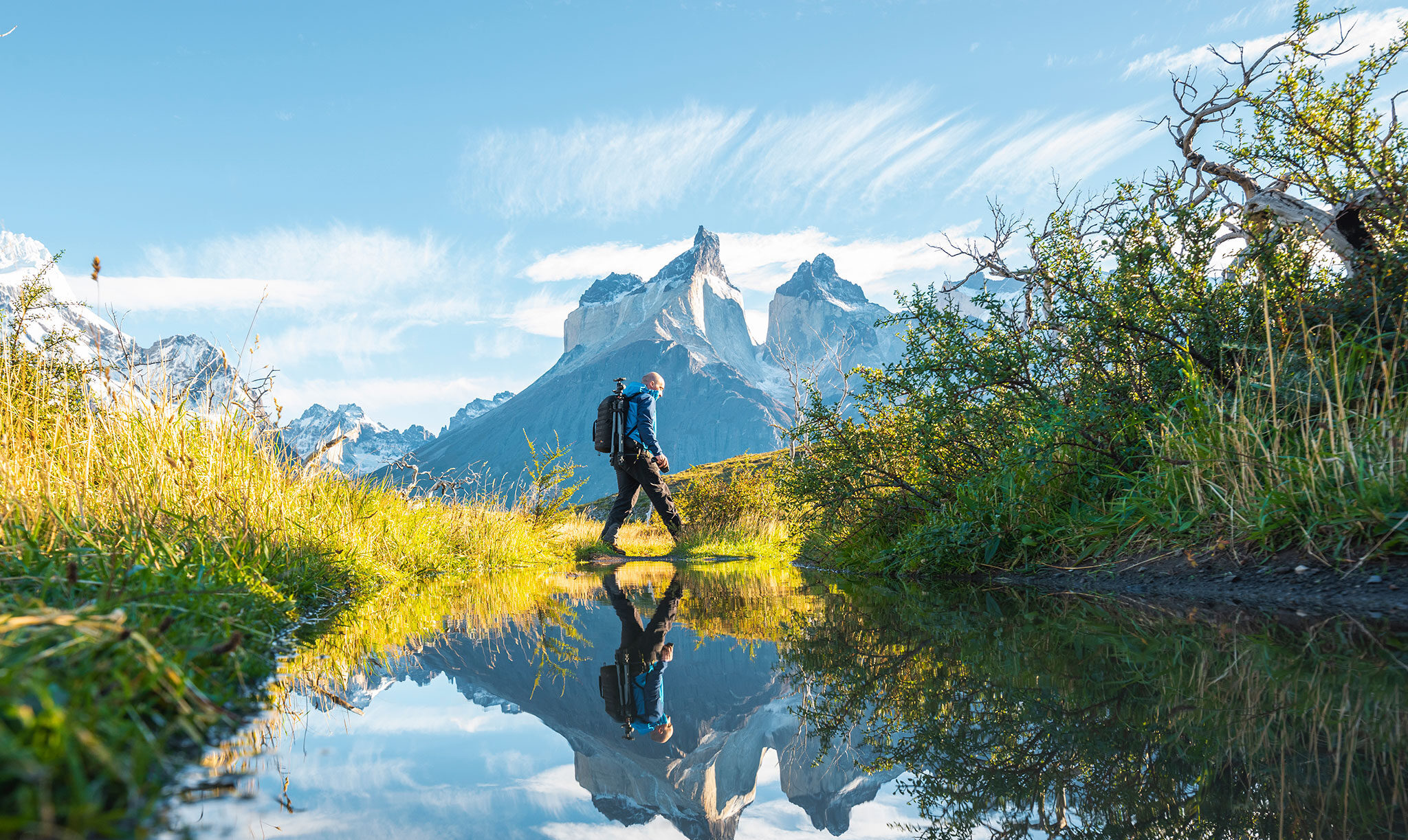 Ein Wanderer mit Fotoausrüstung überquert einen ruhig daliegenden Teich, während in der Ferne scharfe Berggipfel in den Himmel ragen.